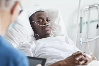 An image of a patient in a hospital bed looking at his doctor with a handheld device in his hand.